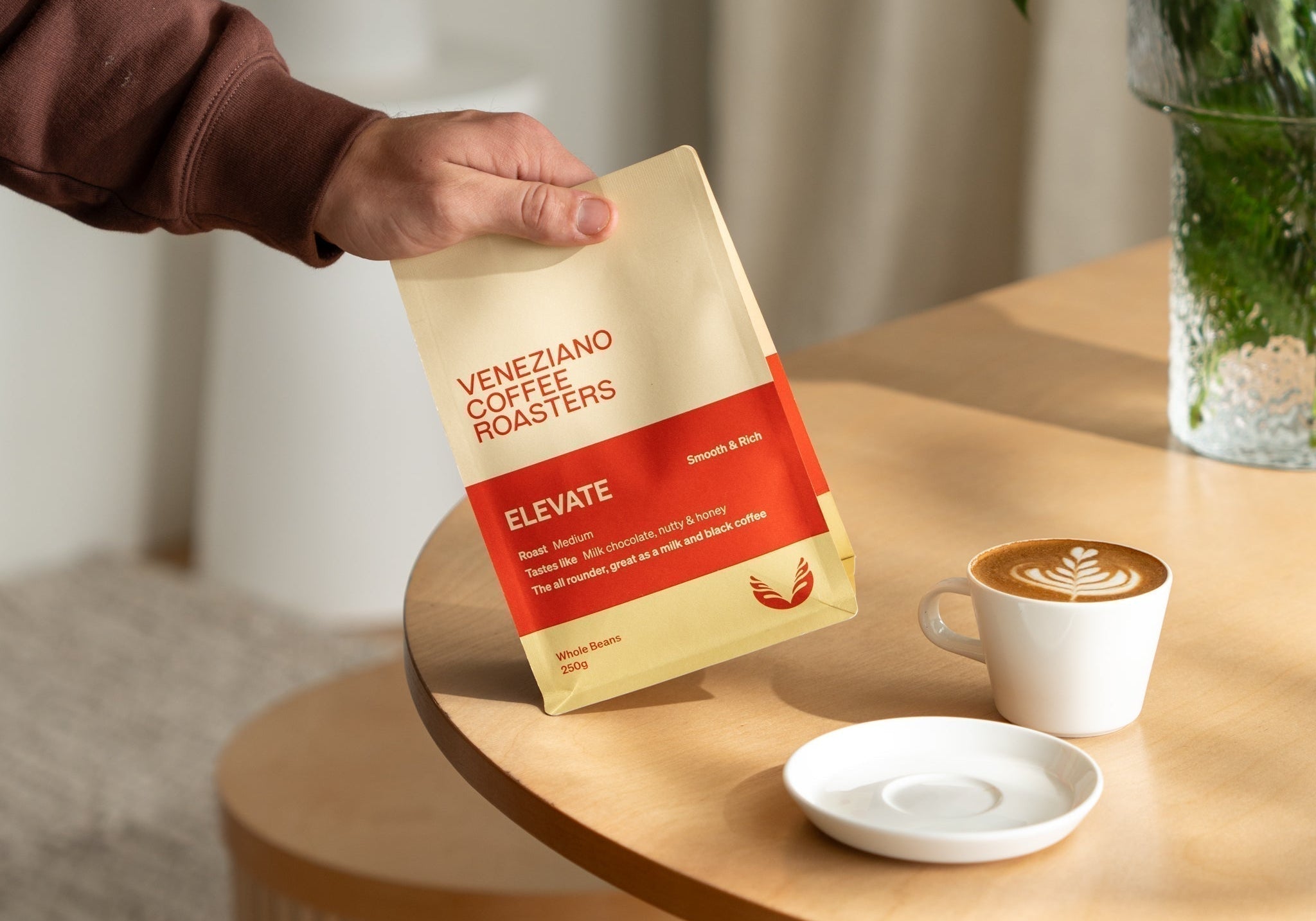 Hand holding a bag of Veneziano's Elevate blend next to a cup of coffee with latte art.