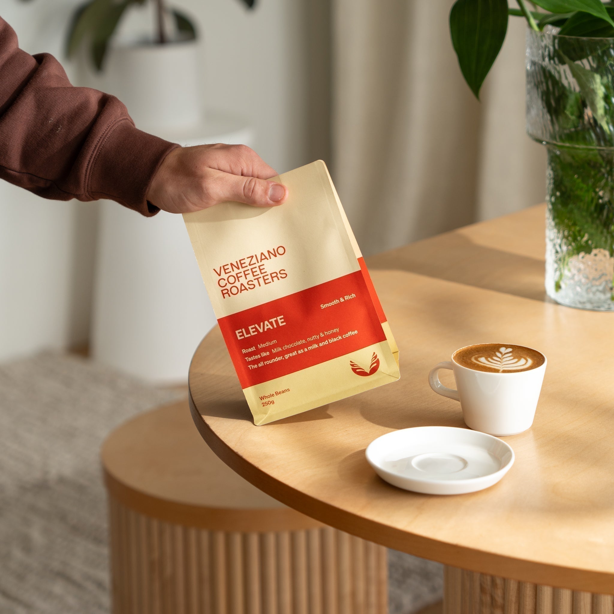 Hand holding a bag of Veneziano's Elevate blend next to a cup of coffee with latte art.