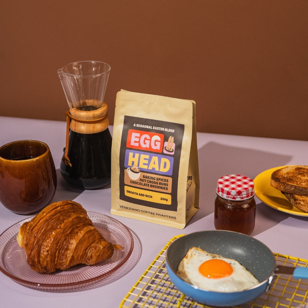 Breakfast setup with coffee, croissant, fried egg, and 'Egg Head' coffee bag on a table.