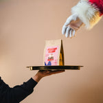 Hand holding a tray with a coffee bag labeled 'Festive Blend' against a beige background