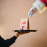 Hand holding a tray with a coffee bag labeled 'Festive Blend' against a beige background