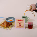 A hand pours coffee from a glass pour-over dripper into a green mug on a tabletop, with a Pinnacle brand packet, a small jar of honey, and breakfast items like a croissant and a fried egg nearby against a plain light background.