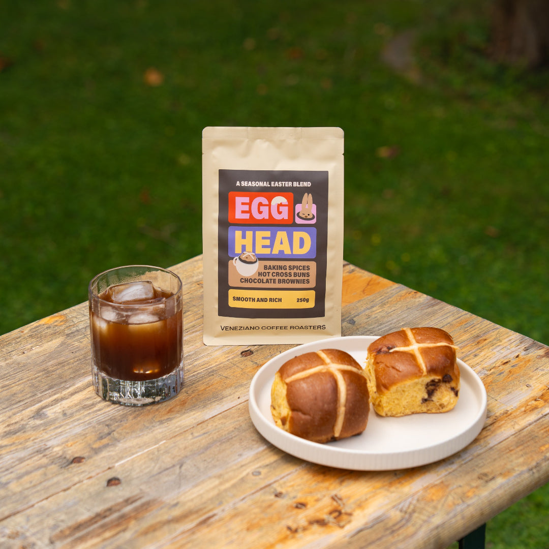 Egg Head coffee package with a glass of iced coffee and hot cross buns on a wooden table outdoors.